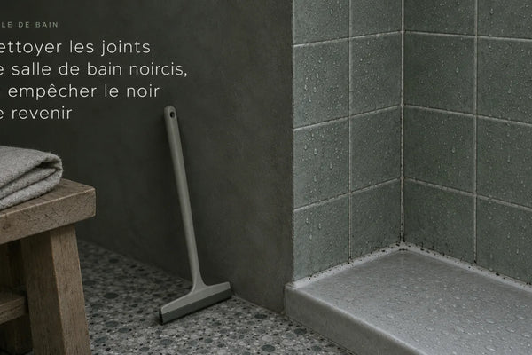Douche aux carreaux gris-vert, joint silicone noirci au pied du receveur, raclette posée contre la paroi, lumière froide du matin